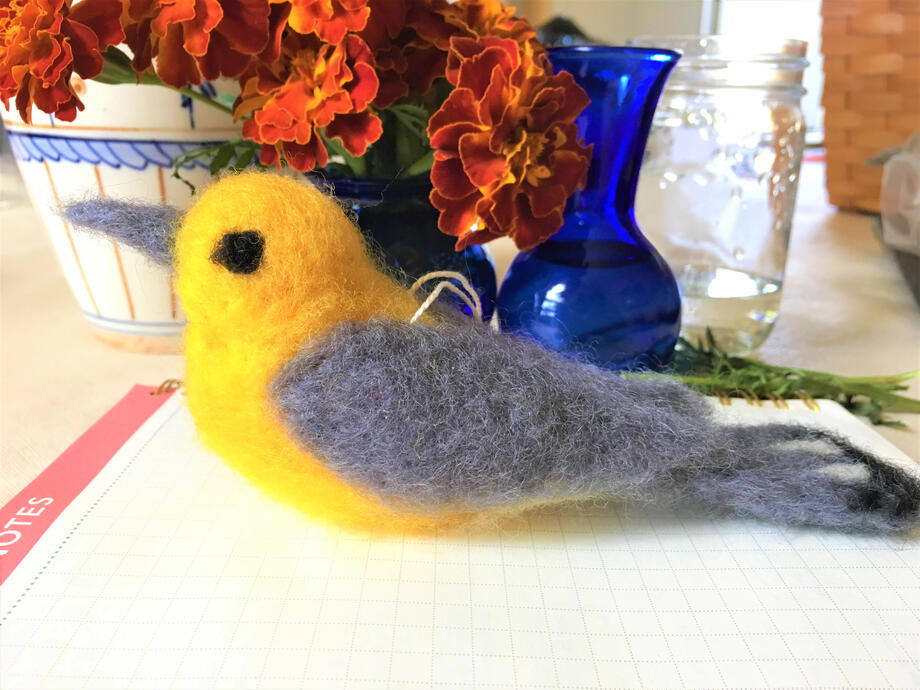 image of a needle felted Prothonotary Warbler, surrounded by decorative objects and flowers